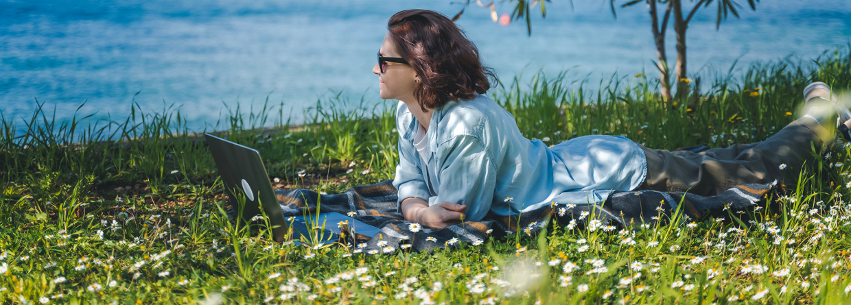 Vrouw is op vakantie en ligt op haar buik in het gras achter haar laptop aan het water