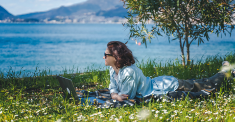 Vrouw is op vakantie en ligt op haar buik in het gras achter haar laptop aan het water